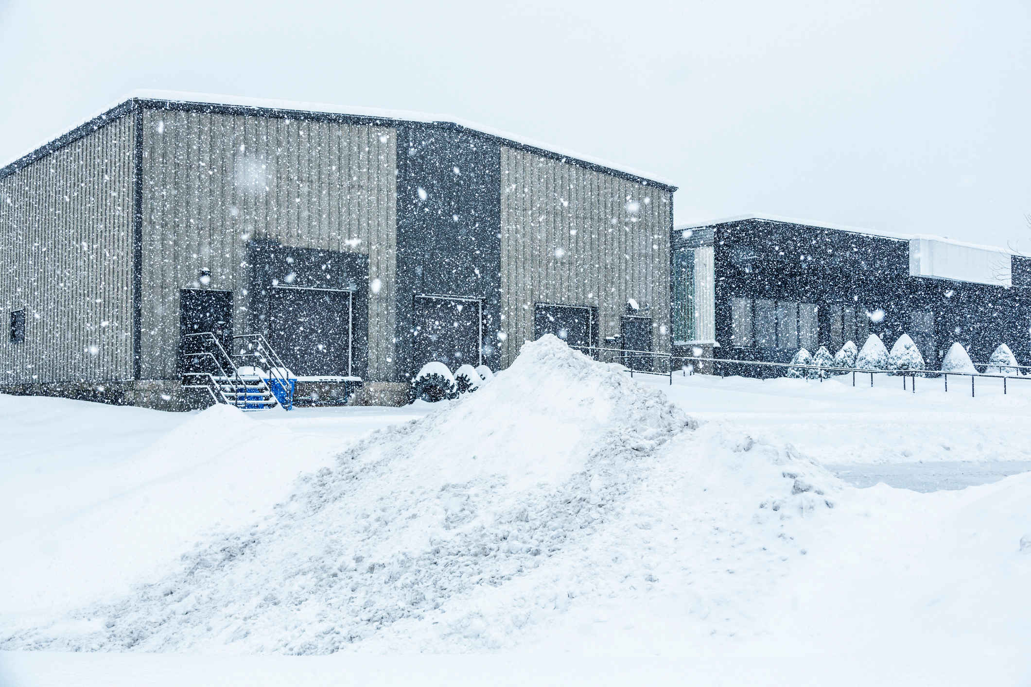 Industrial warehouse loading docks and attached commercial office building behind a heap of plowed and drifting snow at the edge of the parking lot during a blinding blizzard snow storm.